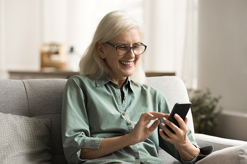 Cheerful blonde old retired woman in glasses using mobile phone, smiling, laughing, sitting on home couch, touching screen, enjoying wireless technology, retirement, leisure