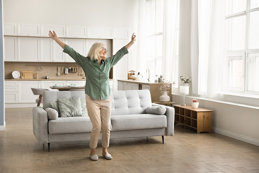 Cheerful carefree elderly retired woman dancing alone in cozy home interior, relaxing in motion, enjoying activity, leisure, retro disco music, good health, healthy joints. Full length shot