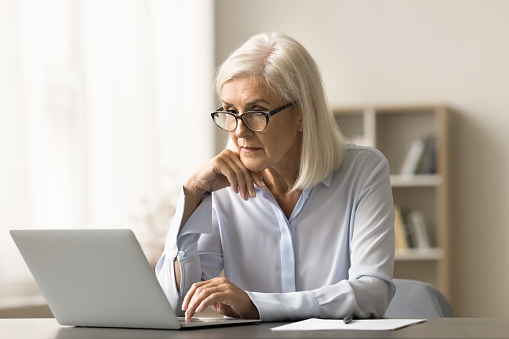 Focused elder business woman in glasses working at laptop computer in home office, using online application, software for job, thinking, making decision, touching chin