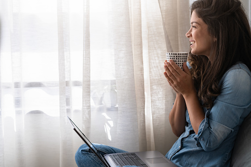 Beautiful smiling young woman sitting at home in cozy window corner, drinking coffee, relaxing, enjoying a morning moment of peace