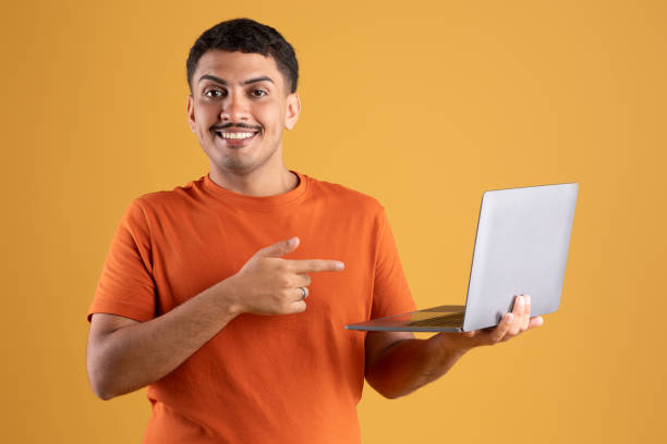 istock Happy latin man pointing at laptop screen and smiling, standing with pc on yellow studio background, reading cool offer 1780537118