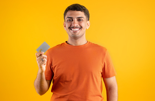 Happy brazilian man holding and showing credit card advertising instant cashless payment, posing over yellow background and smiling at camera