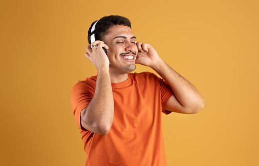 Carefree brazilian man enjoy listening to music with closed eyes and smiling, using modern wireless headphones, standing over orange studio background