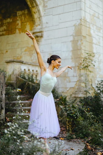 Young graceful woman ballet dancer dressed in professional outfit, shoes and white ballerina skirt dancing with her hands up outside of an abandoned castle. Classic ballet dancer.