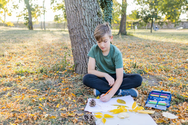 istock Cute schoolboy is doing homework in public park. Creativity for kids is important. Autumn colors and leaves are helping a boy to create wonderful handmade art pieces or crafts. 1780552995