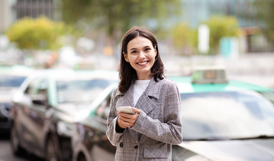 Happy young entrepreneur woman using smart phone, standing in urban area near cars, smiling to camera, messaging and scrolling online news on cellphone gadget, communicating on city street