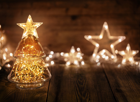 Glass jar in the shape of a Christmas tree with string lights inside on wooden table. Garland in the shape of stars on background. Original decoration for Christmas holidays. Idea of an alternative Christmas tree.