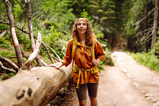 Smiling woman traveler along a forest hiking trail in the mountains against the backdrop of nature. Young woman with backpack traveling outdoors. Hiking, active lifestyle.