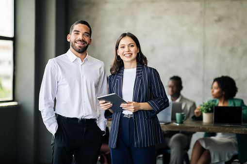 Successful Colleagues. Happy Middle Eastern Businessman And European Businesswoman Standing Holding Digital Tablet, Smiling At Camera In Modern Office During International Business Conference