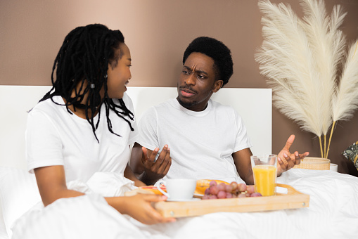 Confused African American dark skinned family couple arguing in bed holding tray with tasty delicious breakfast in bed angry young hasband has quarrel arguments with wife.