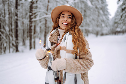 Fashionable woman in a stylish coat and hat holds sparklers in her hands in a snowy forest. A young female tourist in mittens with a New Year's spray bottle. The concept of holidays.