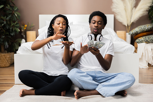 Adorable young stylish African American couple sitting on carpet floor smiling having fun earning a lot of money throwing on floor counting high salary concept.