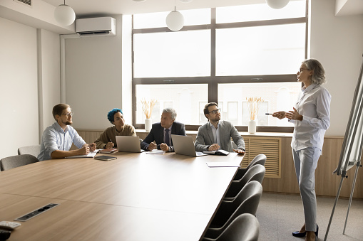 Confident mature female boss speaking to multiethnic team at office meeting table. Businesswoman presenting project to partners. Coach training staff, giving marketing lecture, seminar