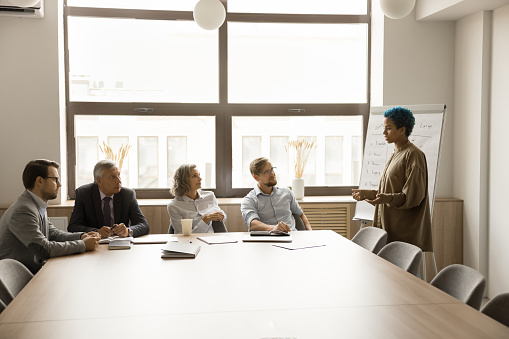 Young African American creative manager girl talking to diverse coworkers, presenting sales report, brainstorming idea, project plan, marketing strategy, speaking at whiteboard on meeting