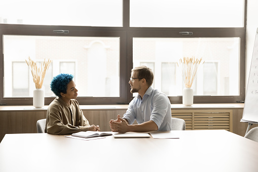 Two positive diverse office friends discussing work business process, teamwork, cooperation, networking in meeting room. Young colleagues talking at large table, collaborating on project