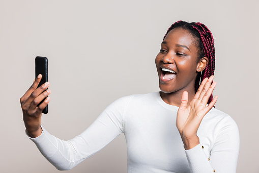 African American woman feeling delighted holds cellphone to participate in a video conference call. The photo captures her in the midst of the video shoot, striking a pose as the process is underway.
