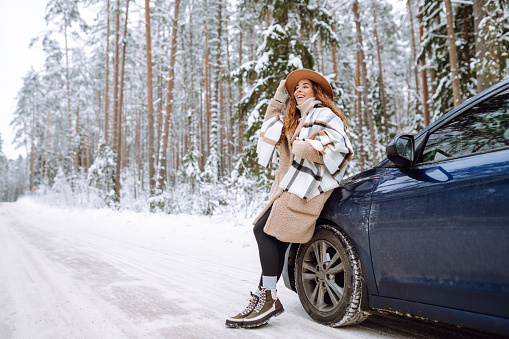 Beautiful young woman in a blue hat and scarf stands near a car on a snowy forest road. A traveler poses near a car in nature. Concept of car travel, vacation.