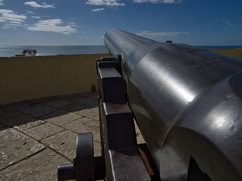 Capture the historic essence of a sun-kissed day with an iron cannon mounted on an iron stand, its wheels positioned towards the vast and endless horizon of the sparkling blue sea. This striking photograph encapsulates the timeless beauty of maritime history, offering a compelling narrative of strength and exploration amidst the serene backdrop of the sunlit sky and the boundless ocean.