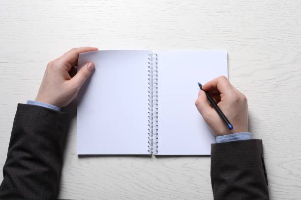 istock Man writing in notebook with pen at white wooden table, top view 1780754867