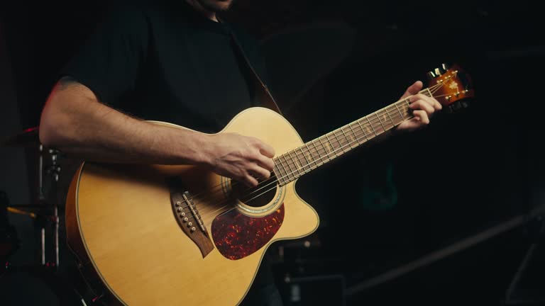 Young musician man sitting and playing acoustic guitar among music equipment in studio close-up. Guitarist plays melody