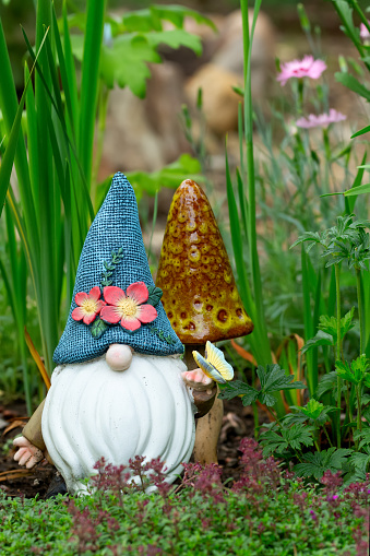Ceramic figurine of a gnome in blue hat holding a little butterfly, standing near a decorative mushroom in a flower bed.