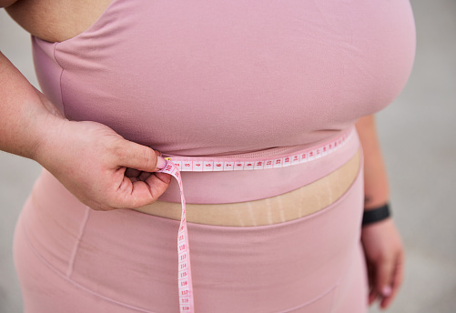 Soft focus of crop anonymous plus size female in pink sportswear measuring waist with tape after weight loss training