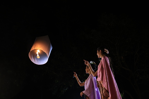 Two beautiful women release a floating lamp into the night sky, symbolizing letting go of problems and the hope for the best on Loi Krathong night.  Embrace of Light concept