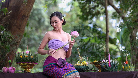 Portrait of a Charming woman in a traditional Thai purple dress and holding a lotus flower in her hand against a forest garden  background on Loi Krathong day