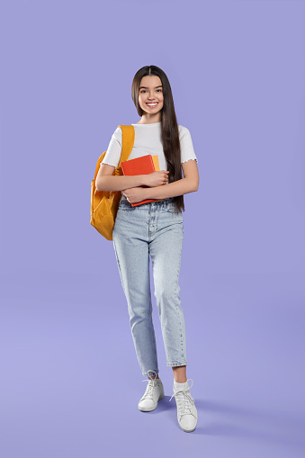 Happy teenage girl with backpack and textbooks on violet background