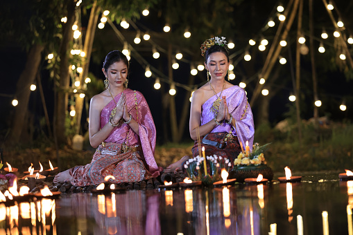 Two beautiful Asian young females in Thai ancient dressing hold and make a wish with Kratong flower floating basket in Loi Kratong festival at night against the yellow  lights backgrounds