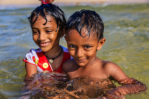 Little smiling child looking at camera while sitting on rocky coast of lake and catching fish with rod in sunlight