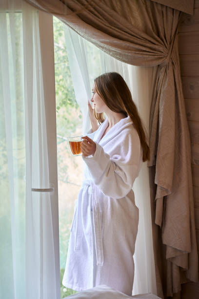 istock Lady wearing bathrobe and holding teacup while staying near window 1780848545