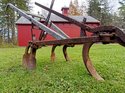 Pittsfield, Massachusetts - USA, October 26, 2023. Old farm plow on green grass with red barn in background in the Berkshires of Massachusetts.