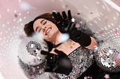 Beautiful woman in gorgeous dress among disco balls under falling confetti in bathtub, above view