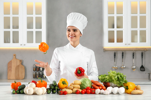 Happy chef throwing bell pepper near fresh vegetables at table in kitchen