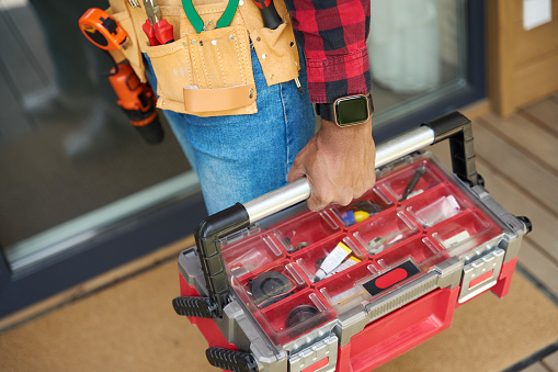 Man with smart watch stands on wooden terrace with a toolbox, he is wearing a red plaid shirt and jeans