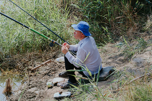 One Caucasian young man in old clothes with a blue hat on his head puts a worm on a fishing rod hook while sitting sideways on the shore of a lake in tall grass on a sunny summer day, side view close-up.