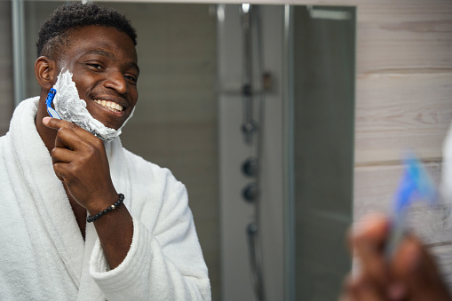 African American guy shaves in front of the mirror in the bathroom, he is wearing a terry robe