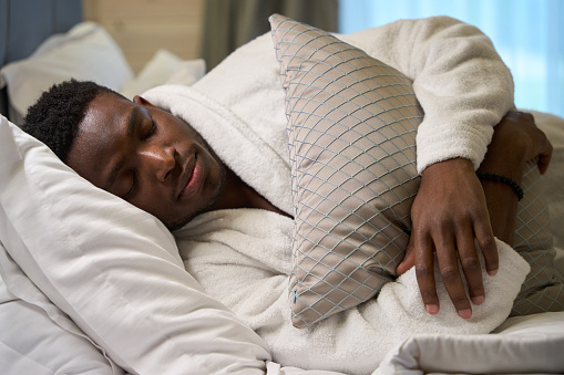 African American guy in a terry robe sleeps sweetly on a large bed, in a modern bedroom interior