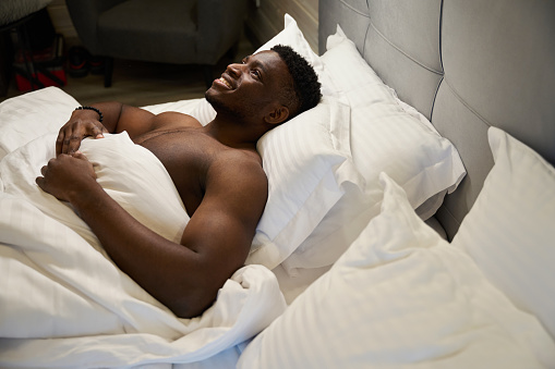 Smiling guy with a naked torso enjoys morning relaxation in a soft bed, he is sitting on soft pillows