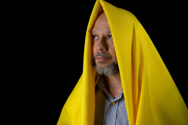 Bald, bearded man with a yellow cloth over his head against black background.