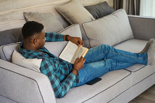 Curly-haired man in casual clothes sits comfortably on the sofa, he enjoys reading a book