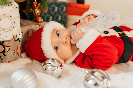 Beautiful little baby boy celebrates Christmas. New Year's holidays. Baby in a Christmas costume Santa's clothes with gifts on fur close to new year tree.