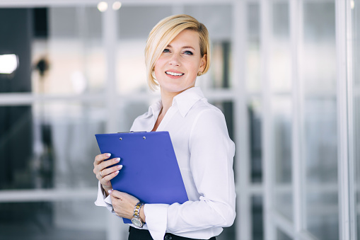 Smiling beautiful business woman holding open folder with documents reading while standing in office