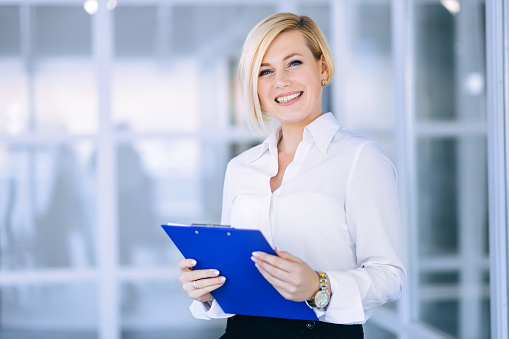 Happy beautiful young business woman standing and holding folder with documents in office