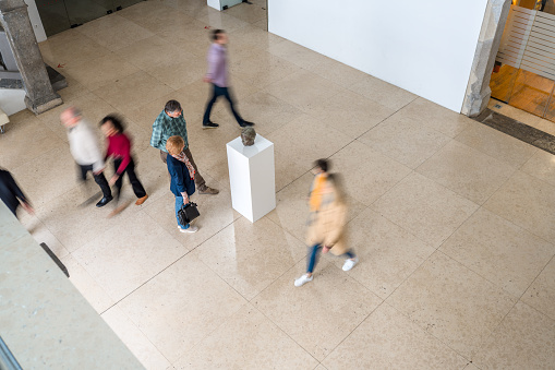 Bird's eye view of the gallery hall with focus on the senior visitors next to a white stand with a bronze sculpture on exhibition. Other people around blurred. Natural and bright light.