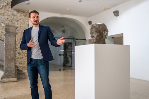 A focused adult black male museum visitor admiring a statue of a man's head . The statue is located on a big white pedestal in the centre of the museum lobby. The man is wearing classy clothes and is standing really close to the statue in order to see better. He is serious as he is looking at the bust. Behind there is a senior couple sitting on the stone bench. The museum is beautiful and bright. It has a stone wall and a stone staircase.