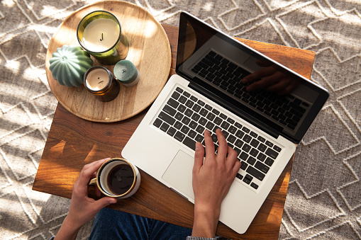 Female hands hold a large mug with coffee at a table in living room next to a laptop