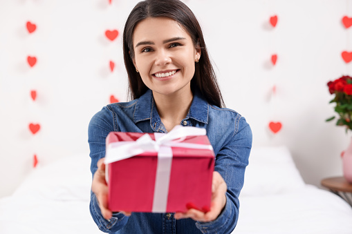 Beautiful young woman with gift box indoors, view from camera. Valentine's day celebration in long distance relationship