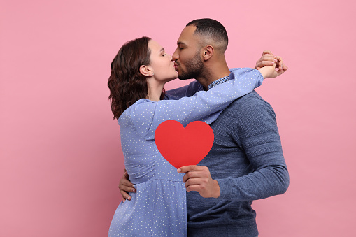 Lovely couple with red paper heart kissing on pink background. Valentine's day celebration
